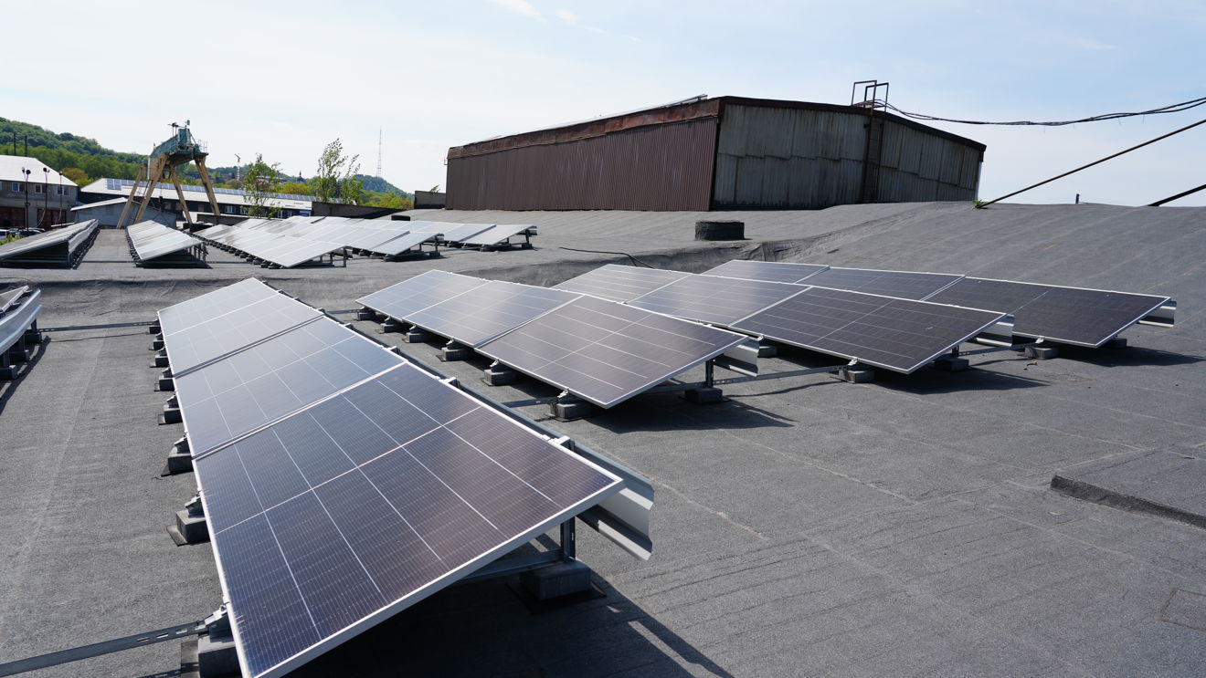 Rows of solar panels are installed on a flat industrial rooftop. Industrial buildings and equipment are visible in the background.