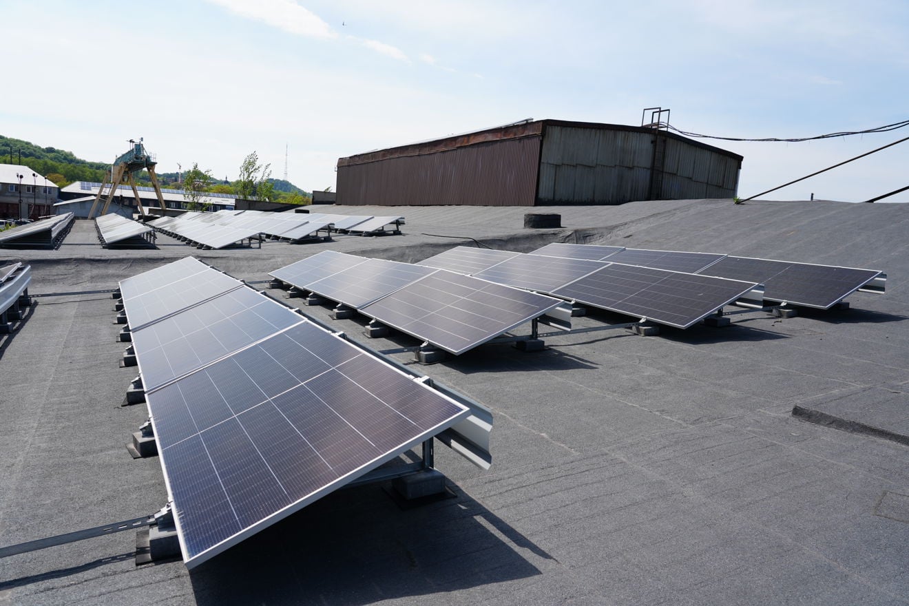 Rows of solar panels are installed on a flat industrial rooftop. Industrial buildings and equipment are visible in the background.