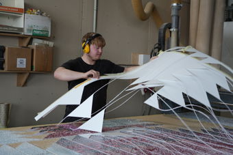 A young man wearing ear defenders works with large white sheet material at a table in a workshop. Cut pieces and strips of material are spread across the work surface in front of him.