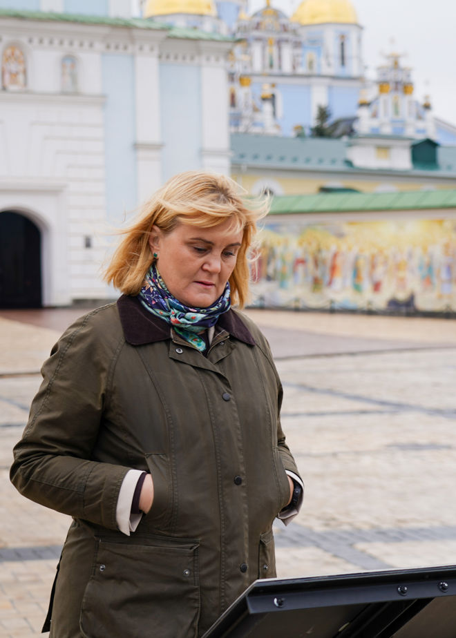 En voksen kvinne med lyst hår og grønn jakke står utendørs foran en informasjonsplakat, med en kirke med gullkupler i bakgrunnen.
