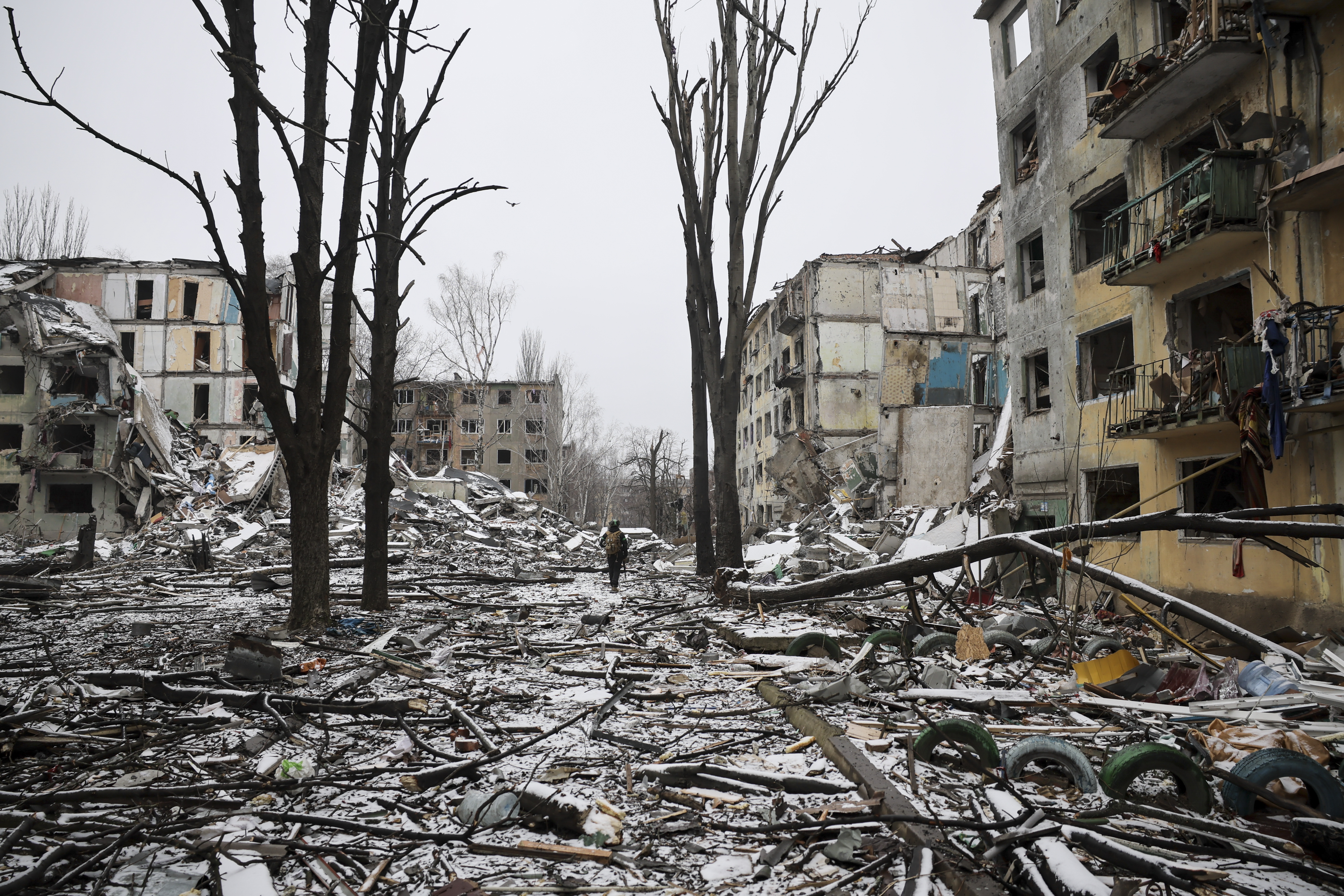 En ødelagt gate med sammenraste bygninger, trær uten blader og snø på bakken; én person går alene blant ruinene.