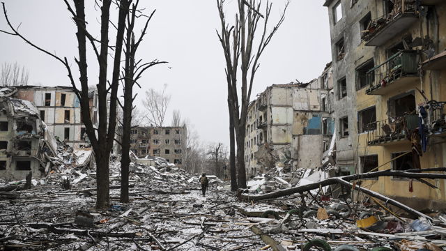 En ødelagt gate med sammenraste bygninger, trær uten blader og snø på bakken; én person går alene blant ruinene.