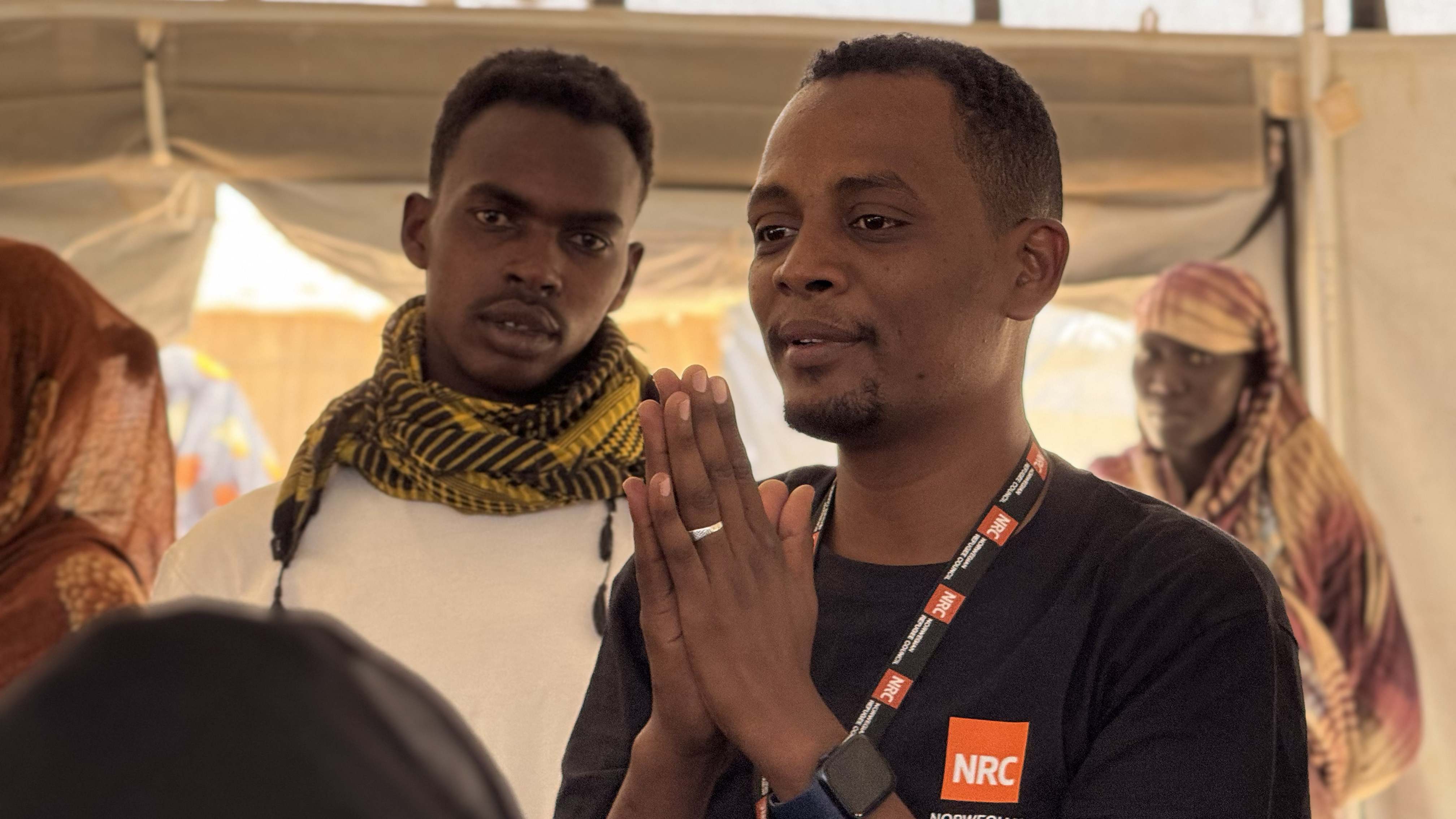 To menn står i et telt, og mannen i forgrunnen snakker med hendene samlet foran seg og har NRC-logo på t-skjorten. Flere personer står i bakgrunnen og lytter.