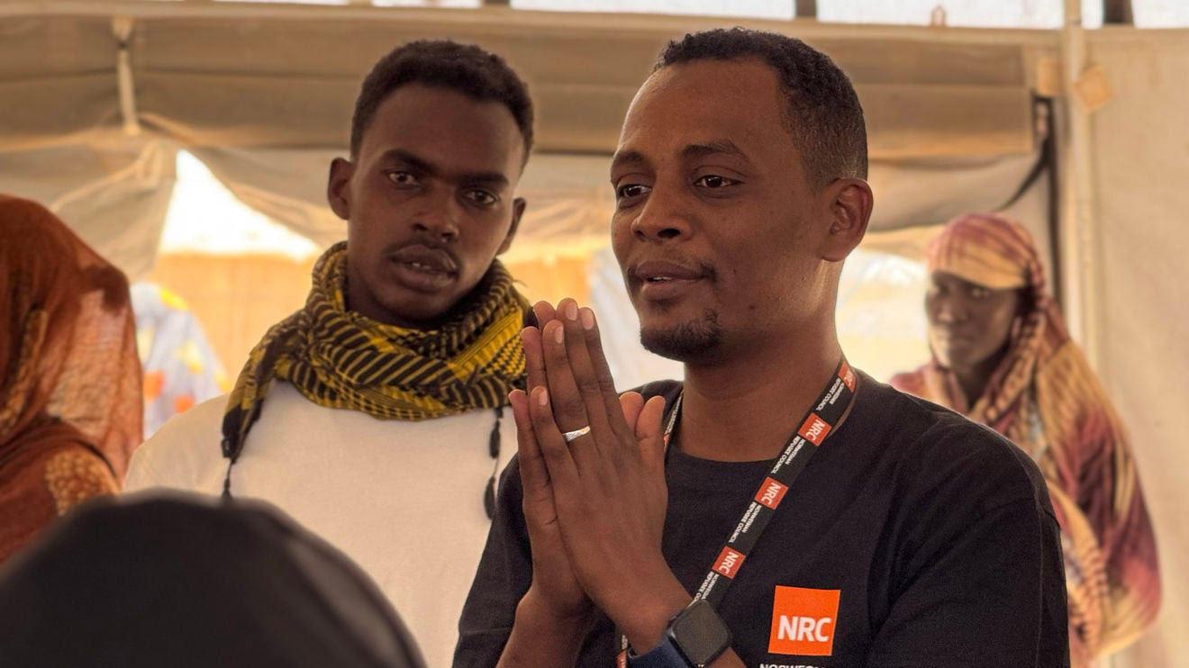 To menn står i et telt, og mannen i forgrunnen snakker med hendene samlet foran seg og har NRC-logo på t-skjorten. Flere personer står i bakgrunnen og lytter.