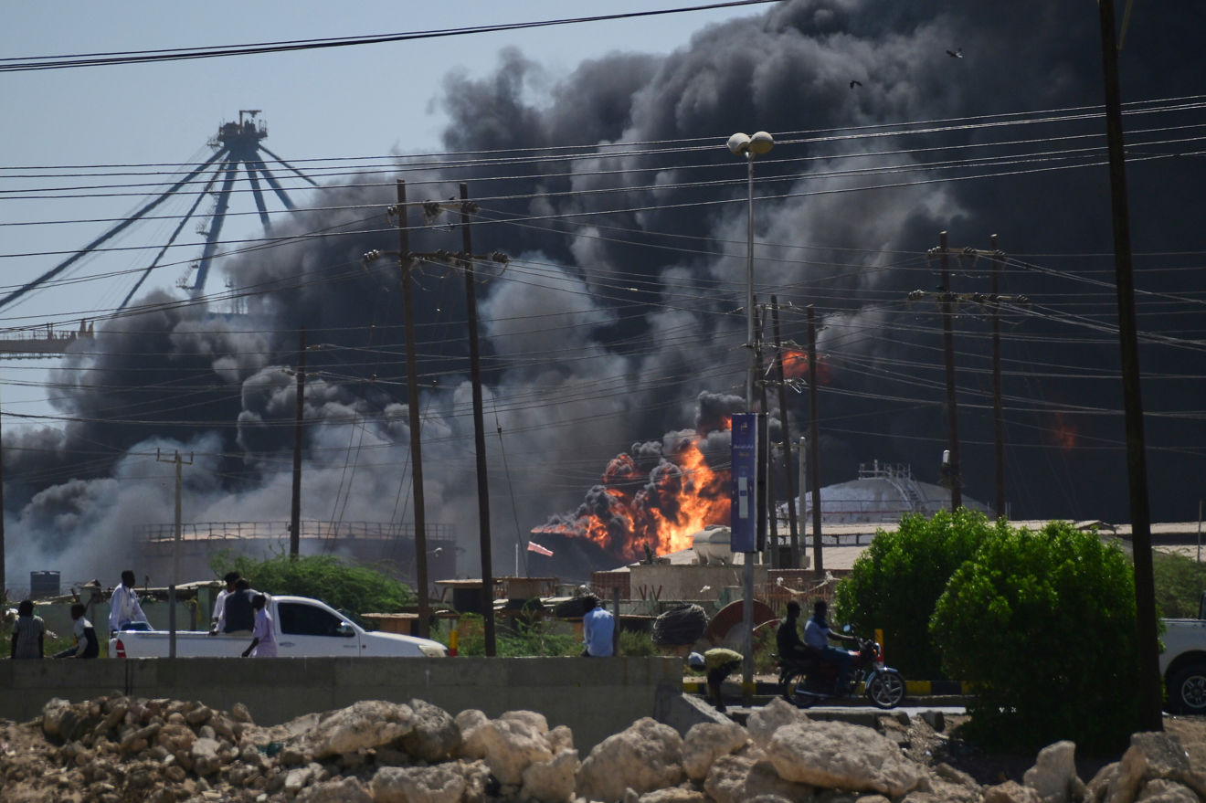 Thick, black clouds of smoke and flames rise from a fire in an industrial area, while people and vehicles move along the road in the foreground.