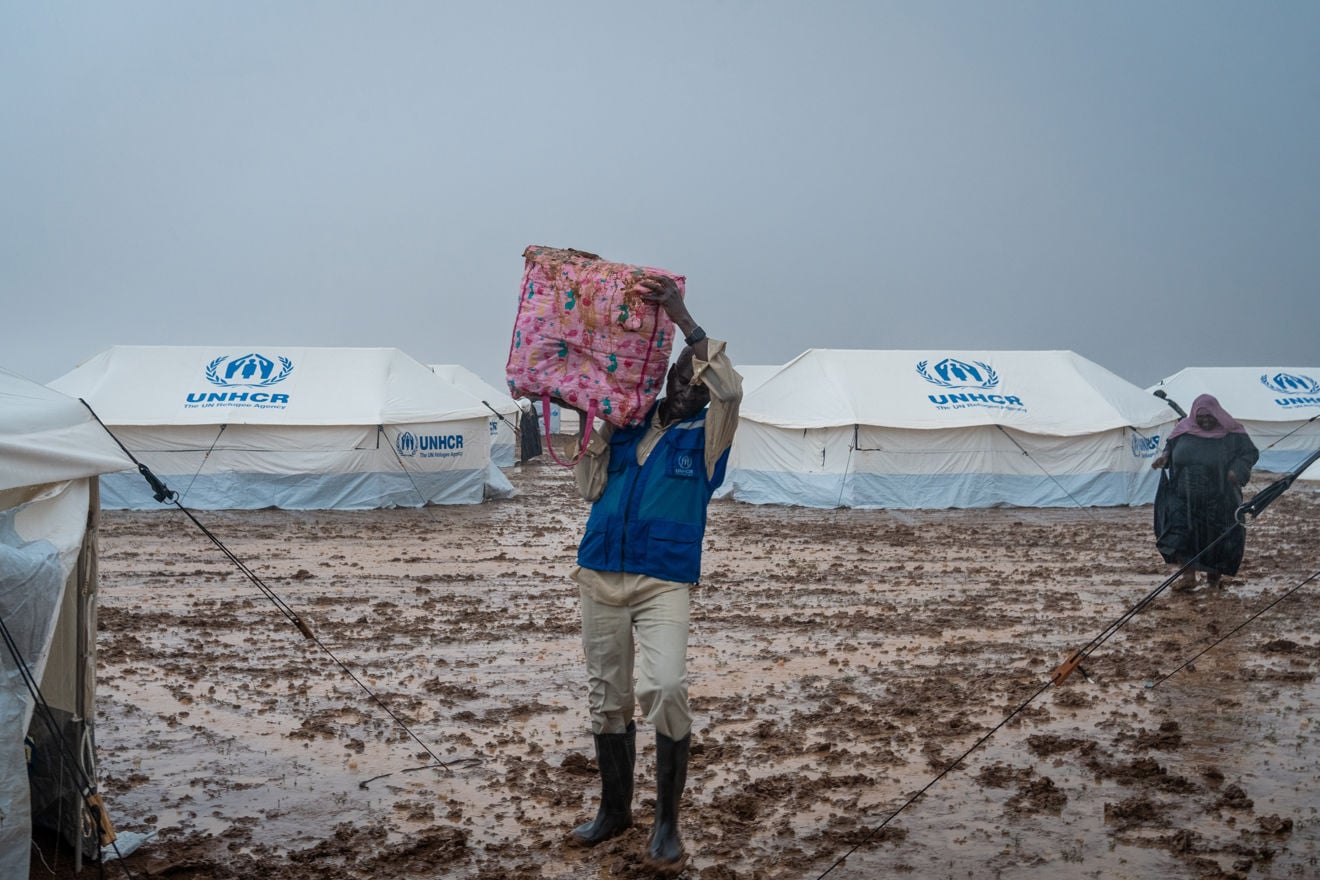 A man wearing boots and a blue vest carries a bag on his head through mud in front of white UNHCR tents in a displacement camp.
