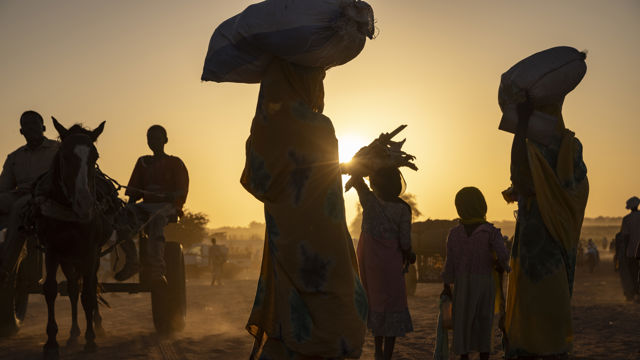 Flere kvinner og barn går på en støvete vei i solnedgang, noen bærer sekker på hodet, mens en hest og kjerre med to menn passerer i bakgrunnen. Bildet er tatt i motlys.