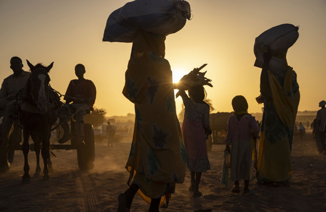 Flere kvinner og barn går på en støvete vei i solnedgang, noen bærer sekker på hodet, mens en hest og kjerre med to menn passerer i bakgrunnen. Bildet er tatt i motlys.