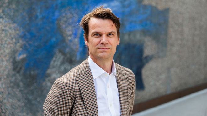 En ung mann i rutete dressjakke og hvit skjorte står utendørs foran en vegg med blått og grått mønster.