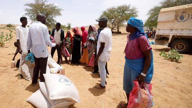A group of adults and children are standing outdoors in line to receive emergency aid, with bags of food and jerrycans in front of them, near a truck marked with the World Food Programme logo.