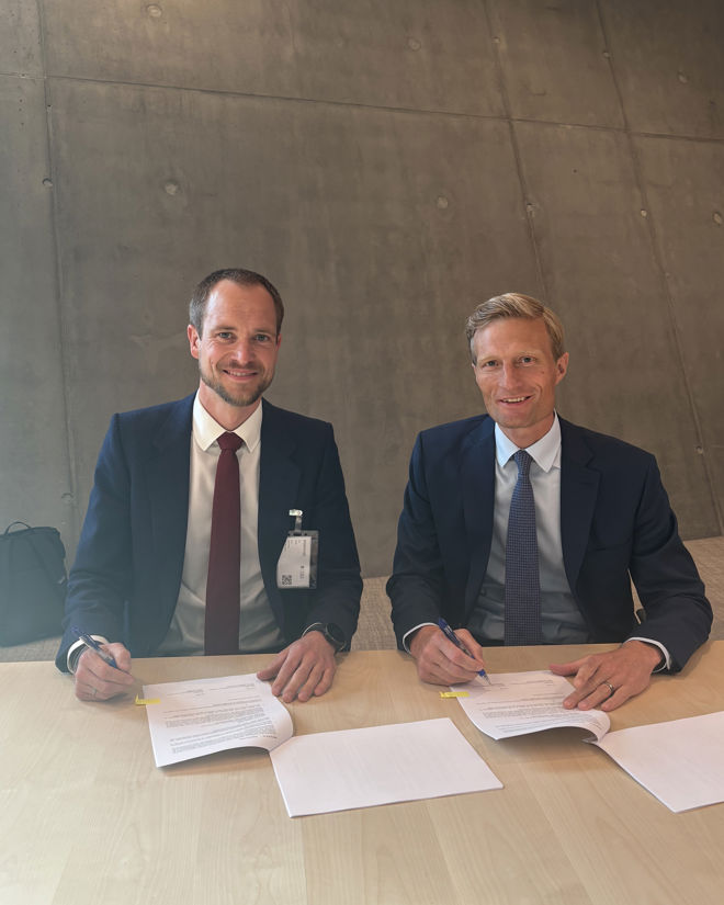  denne  To menn i dress sitter ved et bord og signerer dokumenter.