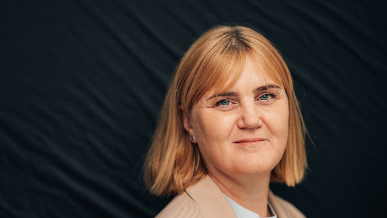 En kvinne med lyst, kort hår står foran en mørk bakgrunn. Hun har på lys blazer og hvit topp og er vendt mot kameraet.
