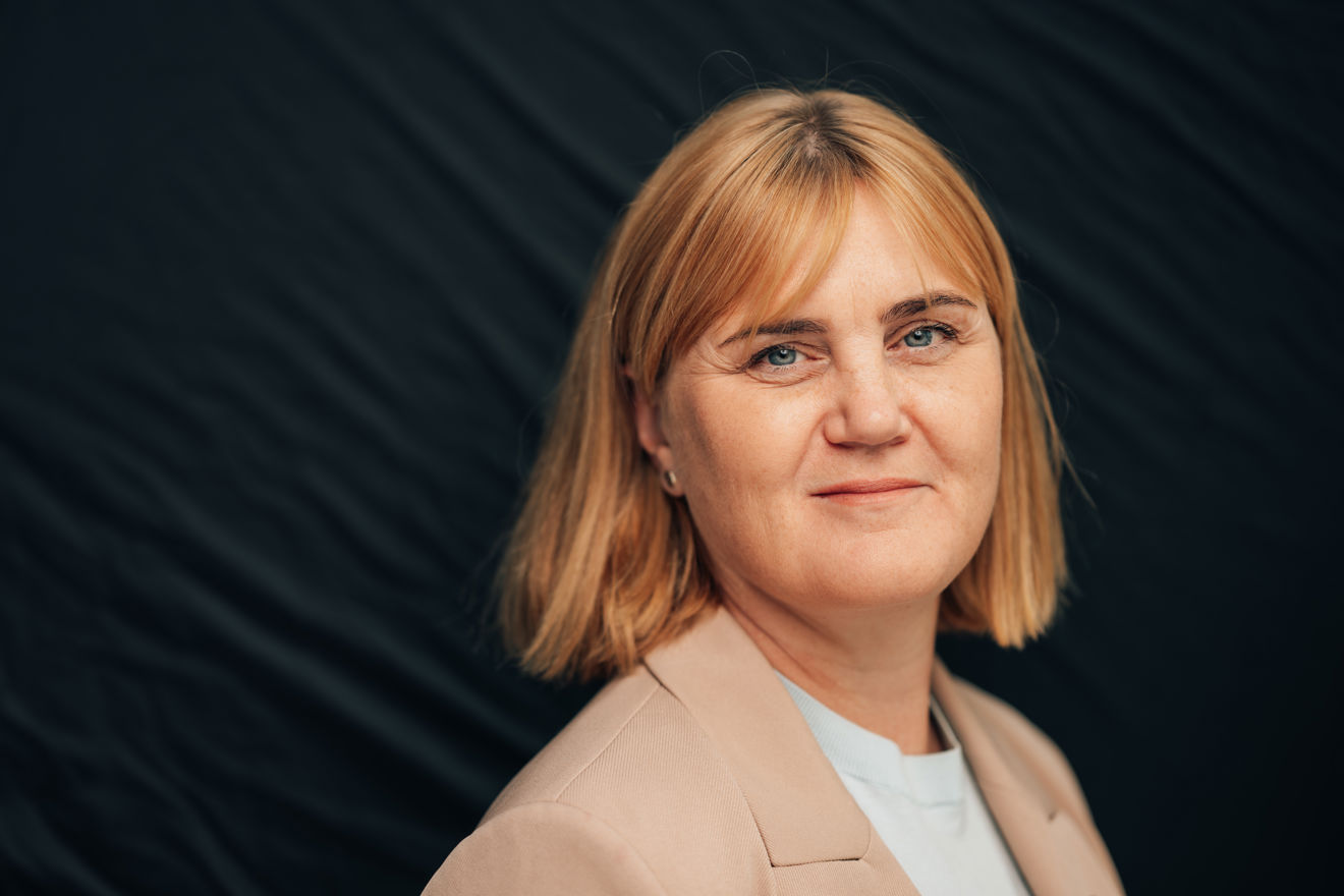 En kvinne med lyst, kort hår står foran en mørk bakgrunn. Hun har på lys blazer og hvit topp og er vendt mot kameraet.