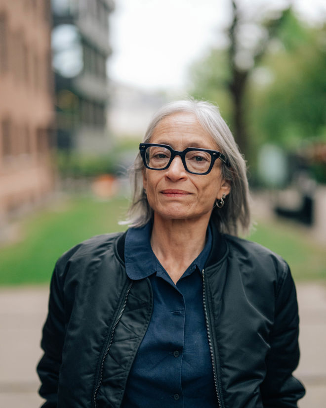 En kvinne står utendørs på en gangvei med hender i jakkelommene. Hun har grått hår og er kledd i mørk jakke og mørk skjorte.