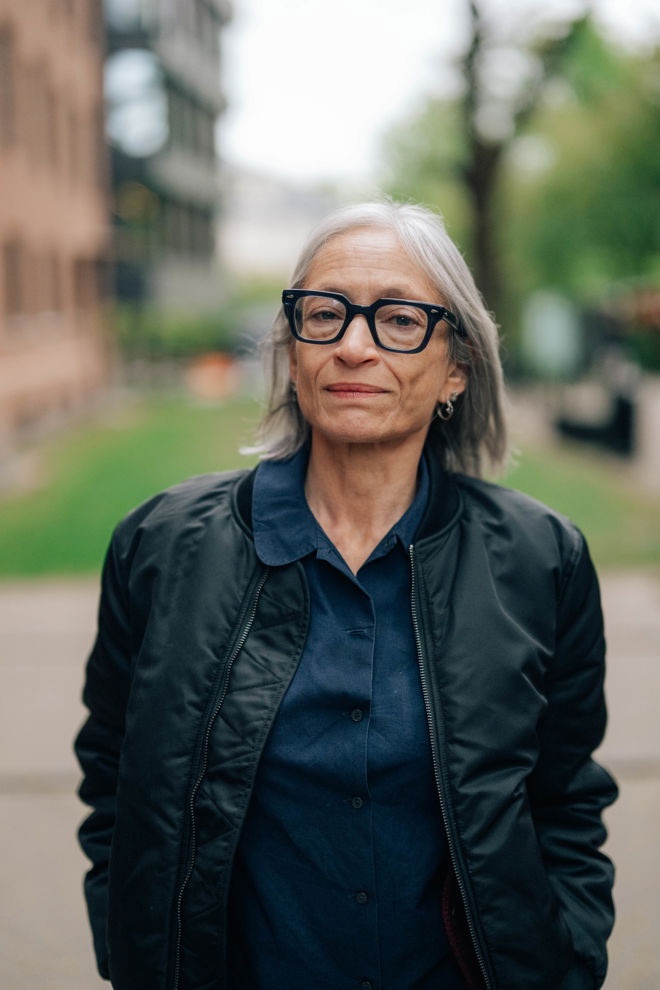 En kvinne står utendørs på en gangvei med hender i jakkelommene. Hun har grått hår og er kledd i mørk jakke og mørk skjorte.