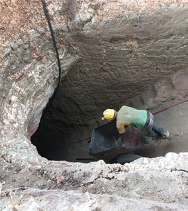 En voksen person med hjelm arbeider nede i en dyp gruve eller sjakt med en trillebår.
