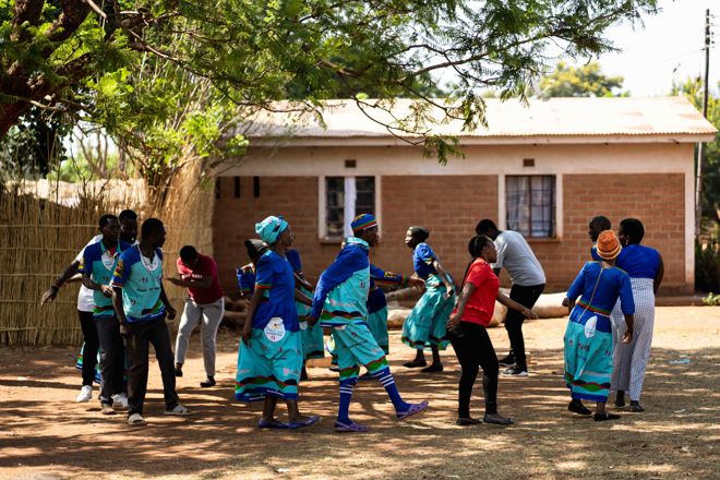 En gruppe unge voksne og voksne danser eller leker sammen utendørs foran et murhus, noen kledd i tradisjonelle blå drakter.