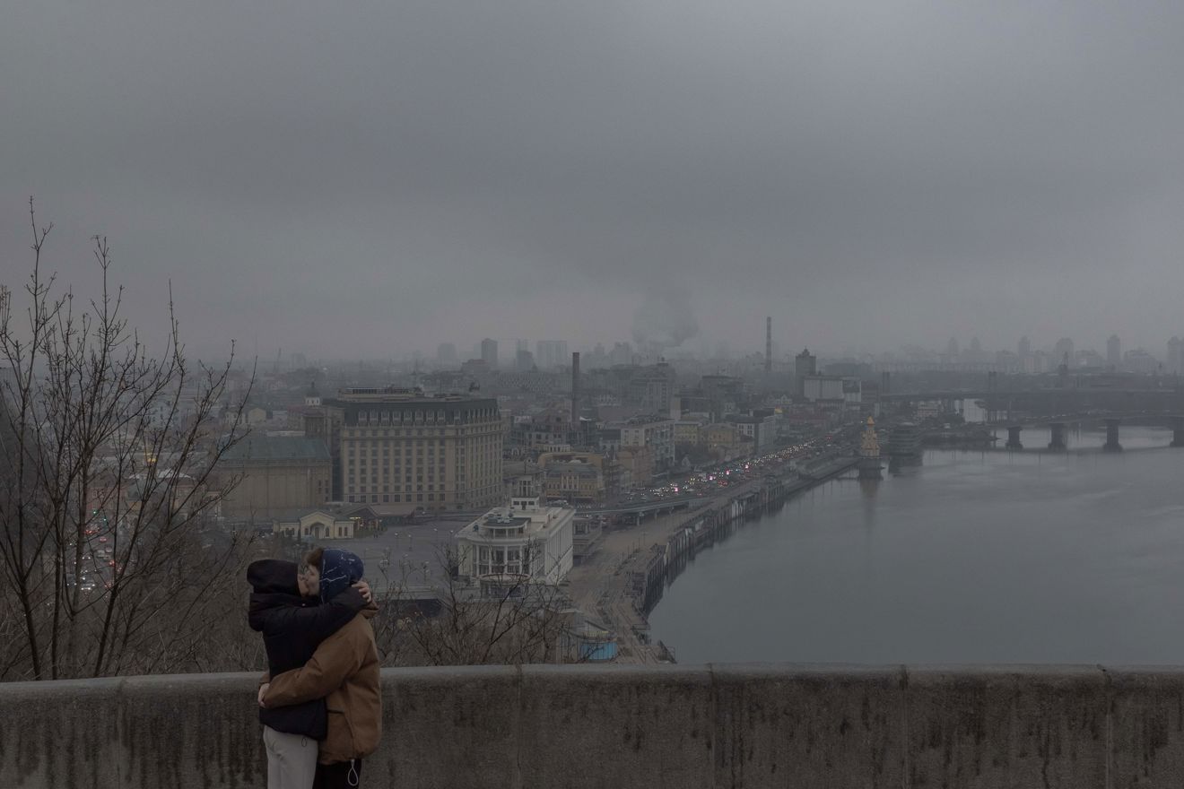 Two people embrace at a railing overlooking a large city and a river on a grey, overcast day.