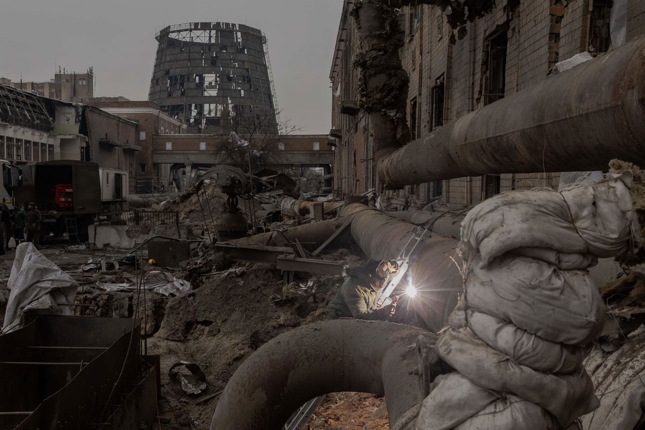 A person welding large pipes in a destroyed industrial area, with ruins and damaged buildings in the background.