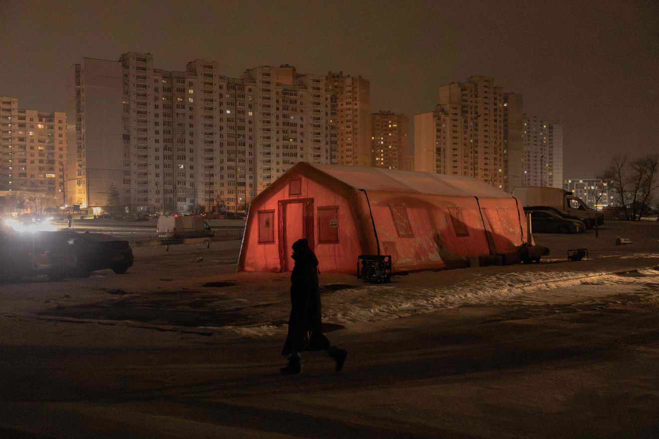 A person walks past a large, reddish tent lit from within, set up in a snow-covered square in front of high-rise apartment buildings at night.
