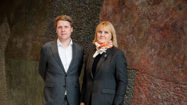 En mann og en kvinne står side om side, begge kledd i mørke dresser foran en steinvegg.