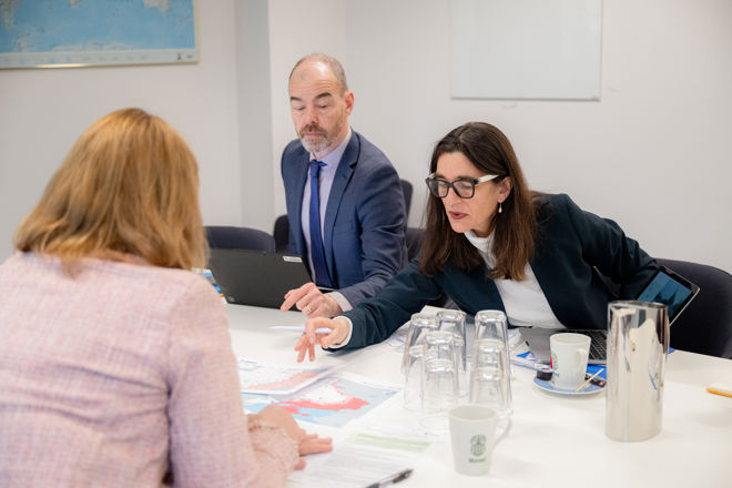 Tre voksne personer sitter rundt et bord og diskuterer dokumenter og kart, med kaffekopper og glass på bordet.