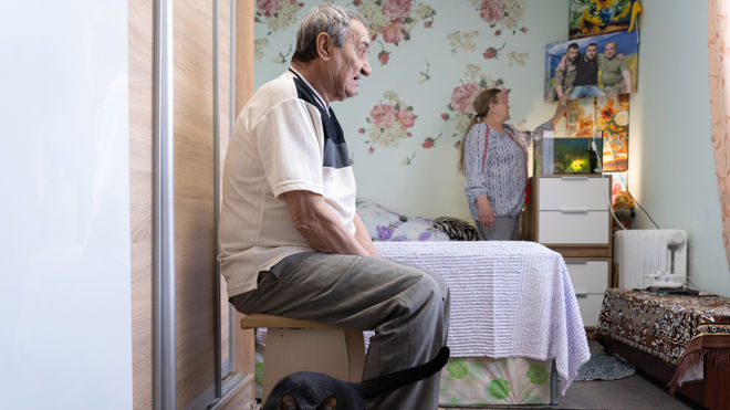 En eldre mann sitter på en krakk i et soverom, mens en kvinne peker på et bilde på veggen; en svart katt er på gulvet.