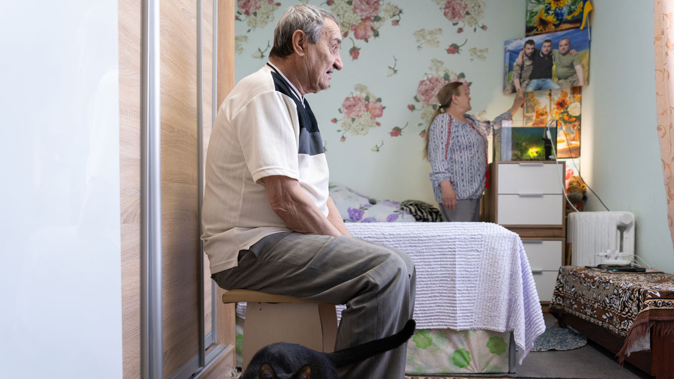 En eldre mann sitter på en krakk i et soverom, mens en kvinne peker på et bilde på veggen; en svart katt er på gulvet.