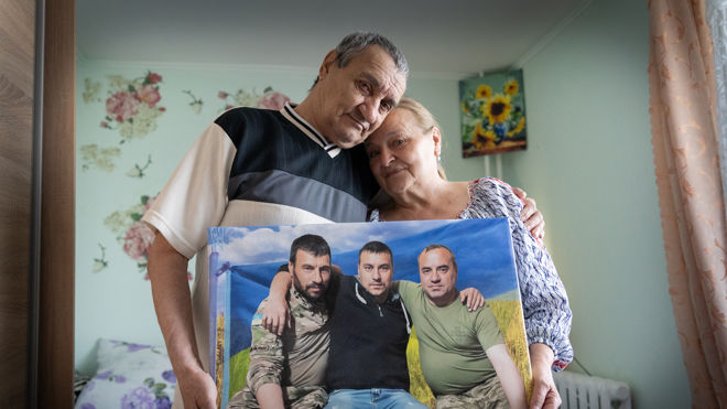 En eldre mann og kvinne står tett sammen og holder et innrammet bilde av tre yngre menn.