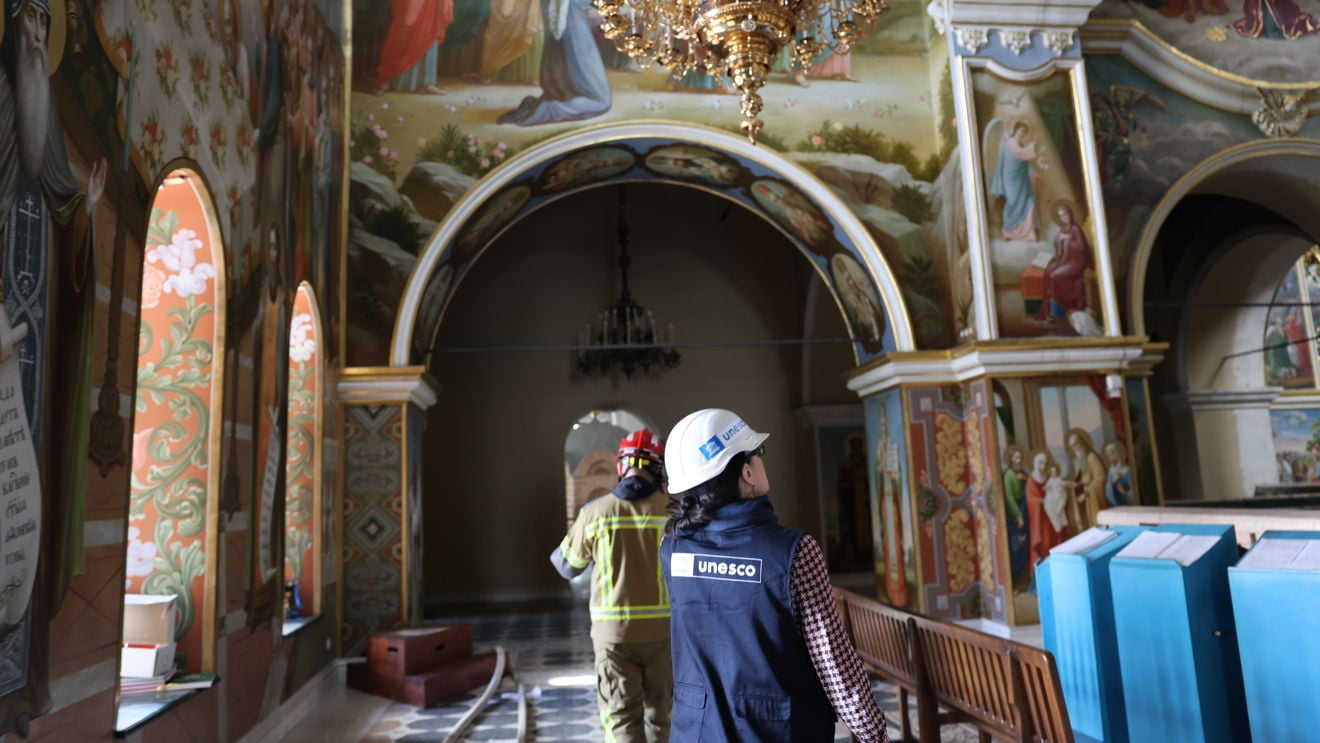 En kvinne med UNESCO-vest og en brannperson går gjennom kirkerommet under veggmalerier og en stor lysekrone. Interiøret har benker, søyler og religiøse motiver på veggene.