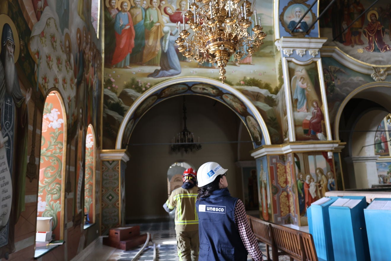 En kvinne med UNESCO-vest og en brannperson går gjennom kirkerommet under veggmalerier og en stor lysekrone. Interiøret har benker, søyler og religiøse motiver på veggene.