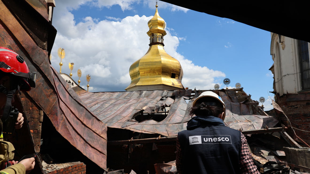 En kvinne med UNESCO-vest og en brannperson står ved et skadet kirketak med hull og sammenrast metall. En gullkuppel er synlig midt i bildet.