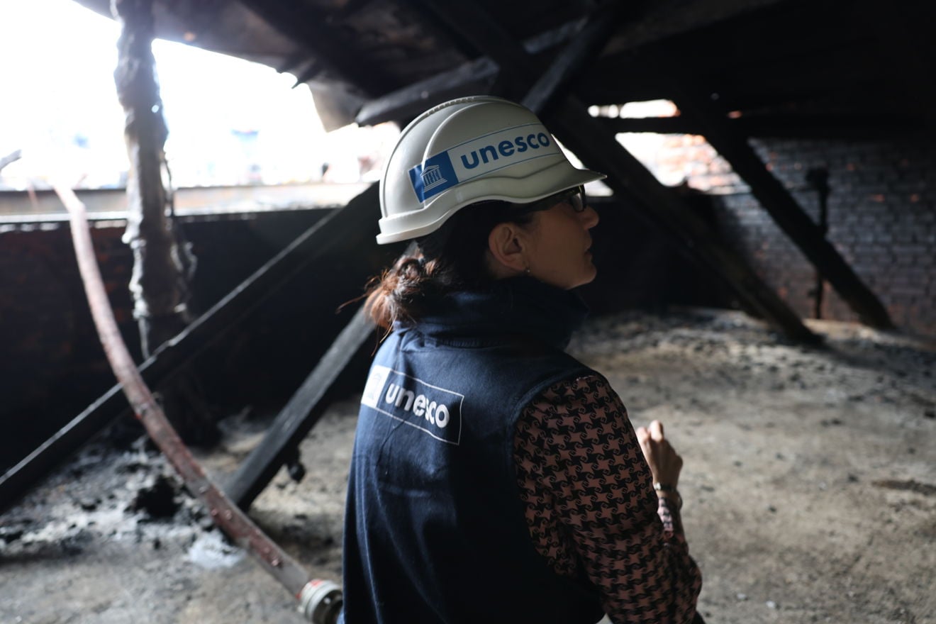 En kvinne med UNESCO-hjelm og vest står inne i et mørkt, brannskadet loftsrom. Forkullede bjelker og brente flater er synlige rundt henne.