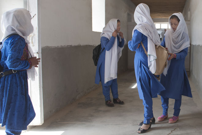 Four young girls wearing blue uniforms and white headscarves are standing and talking together in a school hallway.