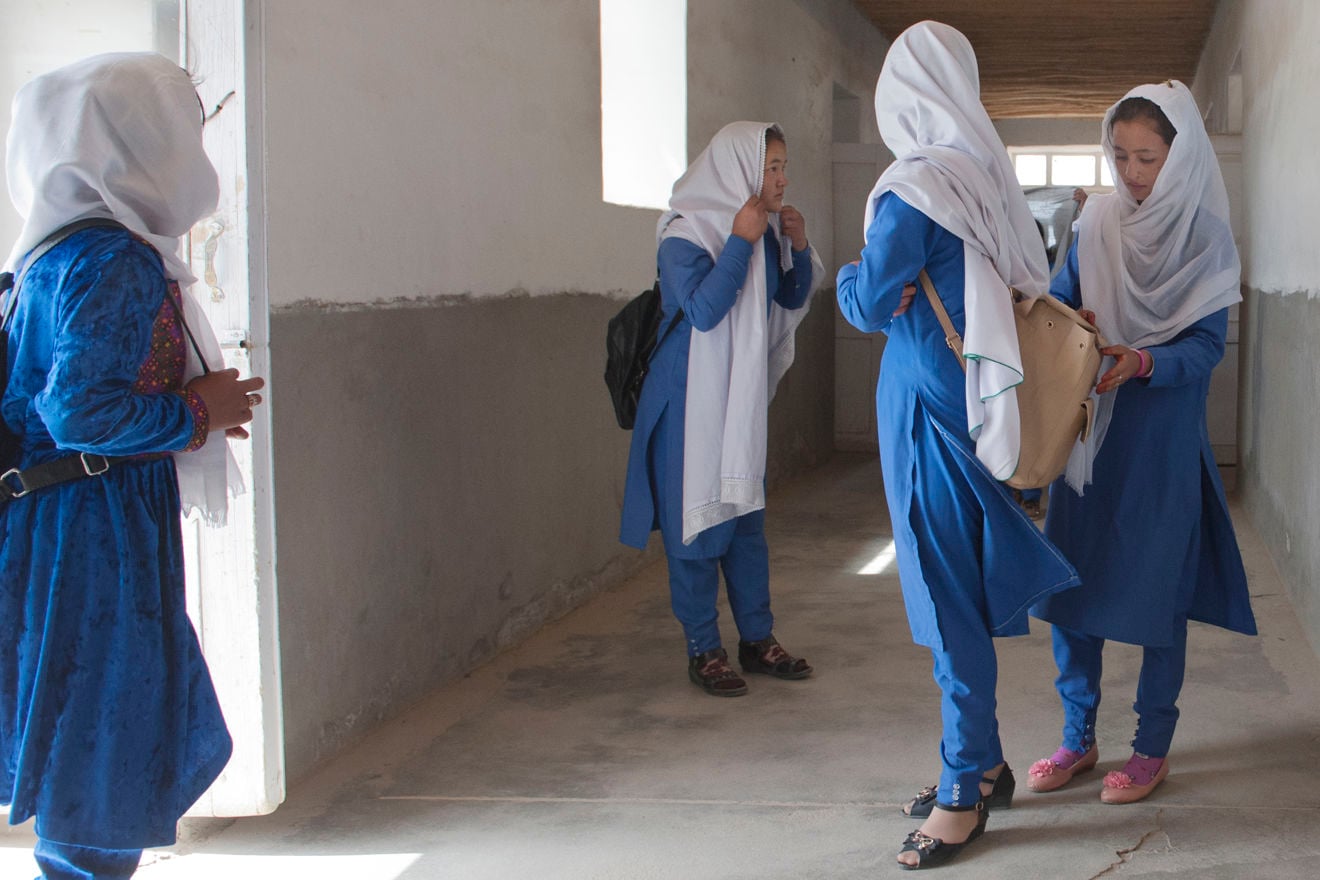 Four young girls wearing blue uniforms and white headscarves are standing and talking together in a school hallway.