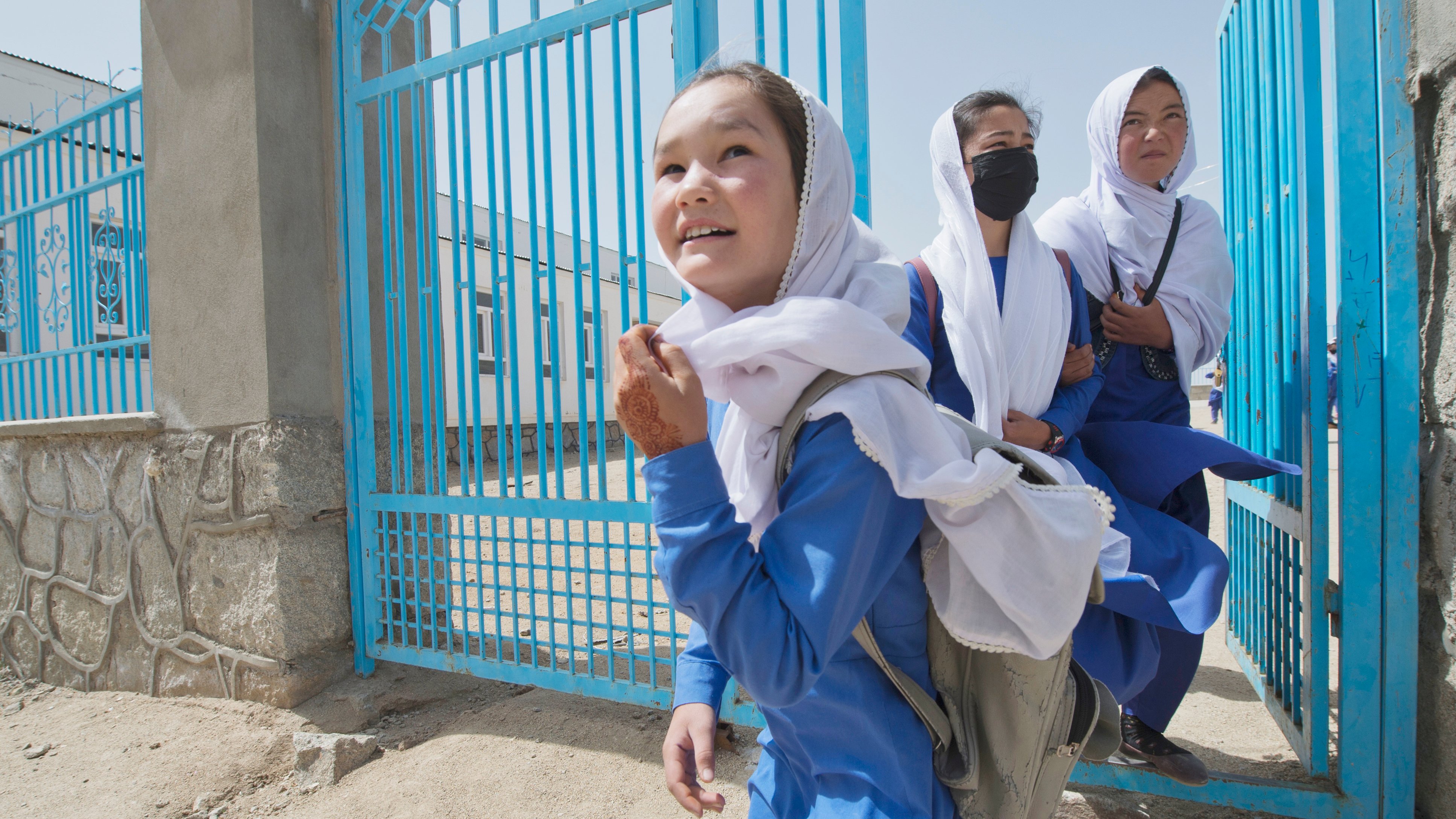 Three young girls wearing blue uniforms and white headscarves are walking out of a blue school gate.
