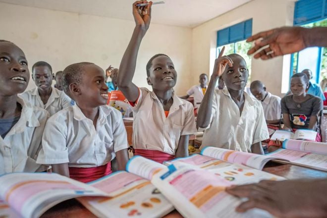 Several young boys and girls in school uniforms are sitting in a classroom, with some raising their hands and textbooks open on their desks.