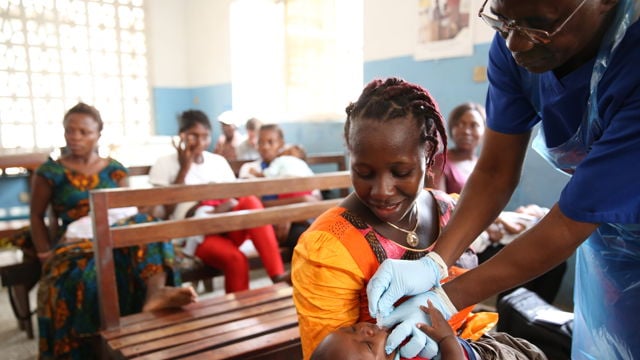 En helsearbeider gir vaksine til en liten gutt som sitter på fanget til en kvinne, med flere voksne og barn sittende på benker i bakgrunnen.