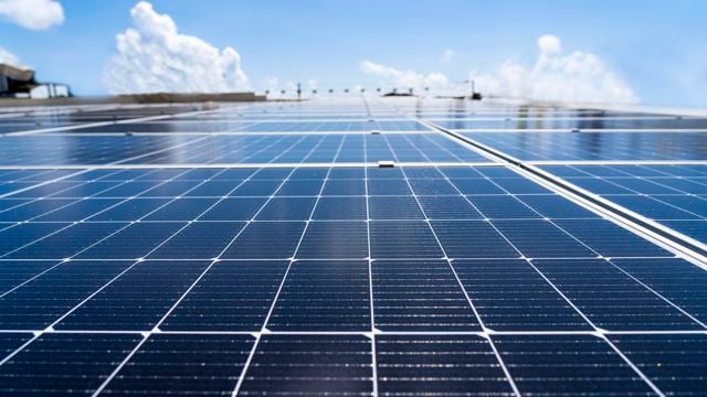 The image shows a close-up of solar panels stretching horizontally across a surface. The panels have a glossy finish that reflects the sunlight. In the background, a clear blue sky with some scattered white clouds can be seen.