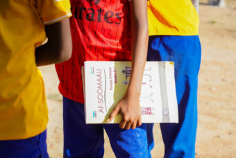 Bildet viser et barn som står ute og holder en bok med tittelen "AF-SOOMAALI", som er på somali. Barnet har på seg rød skjorte og blå bukser. Ved siden av står andre barn i gule overdeler og blå bukser.