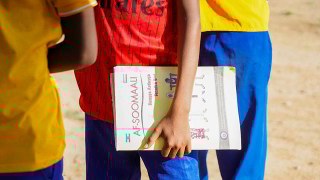 The image shows a person holding a book or booklet titled "AF-SOOMAALI" in their hand. They are wearing a red shirt with a graphic design and blue pants. Another person nearby is wearing a yellow shirt. The setting appears to be outdoors, with a sandy ground visible, suggesting a casual or school environment.