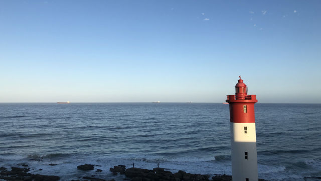 The image depicts a scenic ocean view with a red and white lighthouse positioned on the right side. The lighthouse stands beside a rocky shoreline, overlooking the vast expanse of the sea. The sky is clear and blue, adding to the tranquil atmosphere. In the distance, ships can be seen on the horizon, emphasizing the maritime setting.