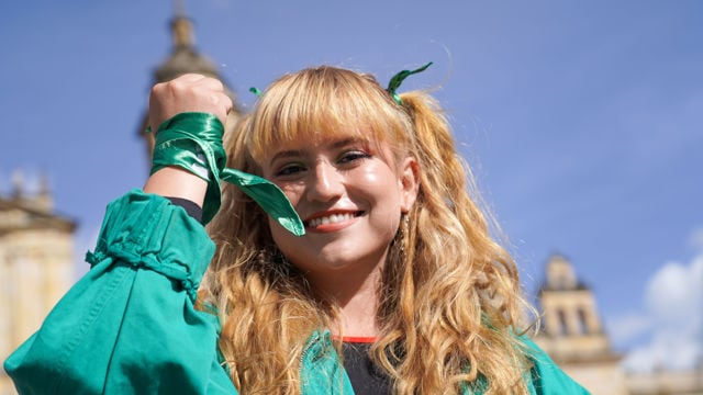 The image shows a person with long, curly blonde hair tied with green ribbons. They are wearing a green jacket and have a green cloth wrapped around their raised fist. The background features a clear blue sky and parts of a historical building, suggesting an outdoor setting, possibly during a demonstration or protest.