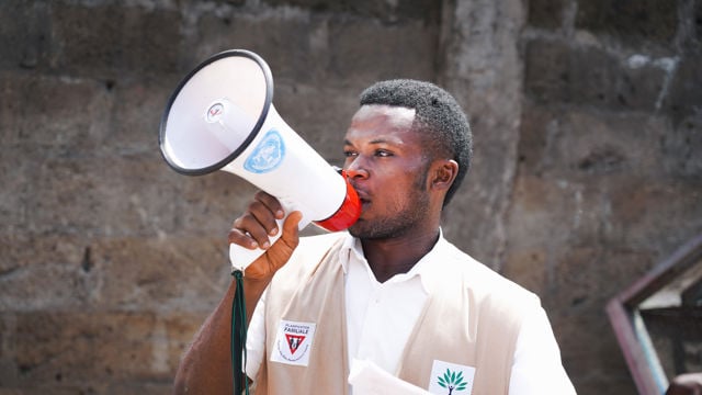 An adult man holds a megaphone with a UN logo in front of a stone wall.