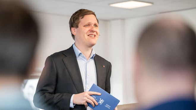 Bildet viser en person kledd i en formell mørk dress og en lys blå skjorte, som holder et blått hefte med et emblem eller våpenskjold på