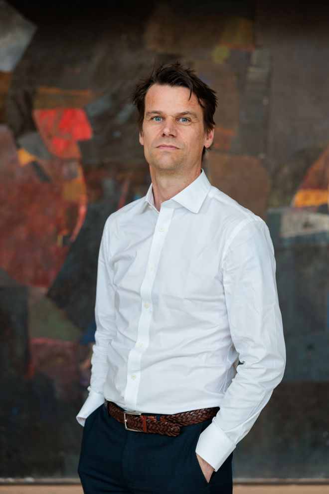 En mann med hvit skjorte og mørke bukser står foran en fargerik bakgrunn med hendene i lommene.