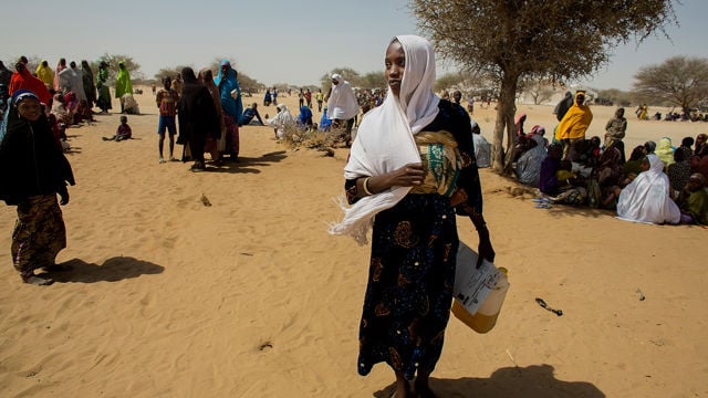 A woman stands in the foreground of a sunlit, sandy landscape, holding a plastic container and some papers. She wears a dark patterned dress and a white scarf draped over her head and shoulders.