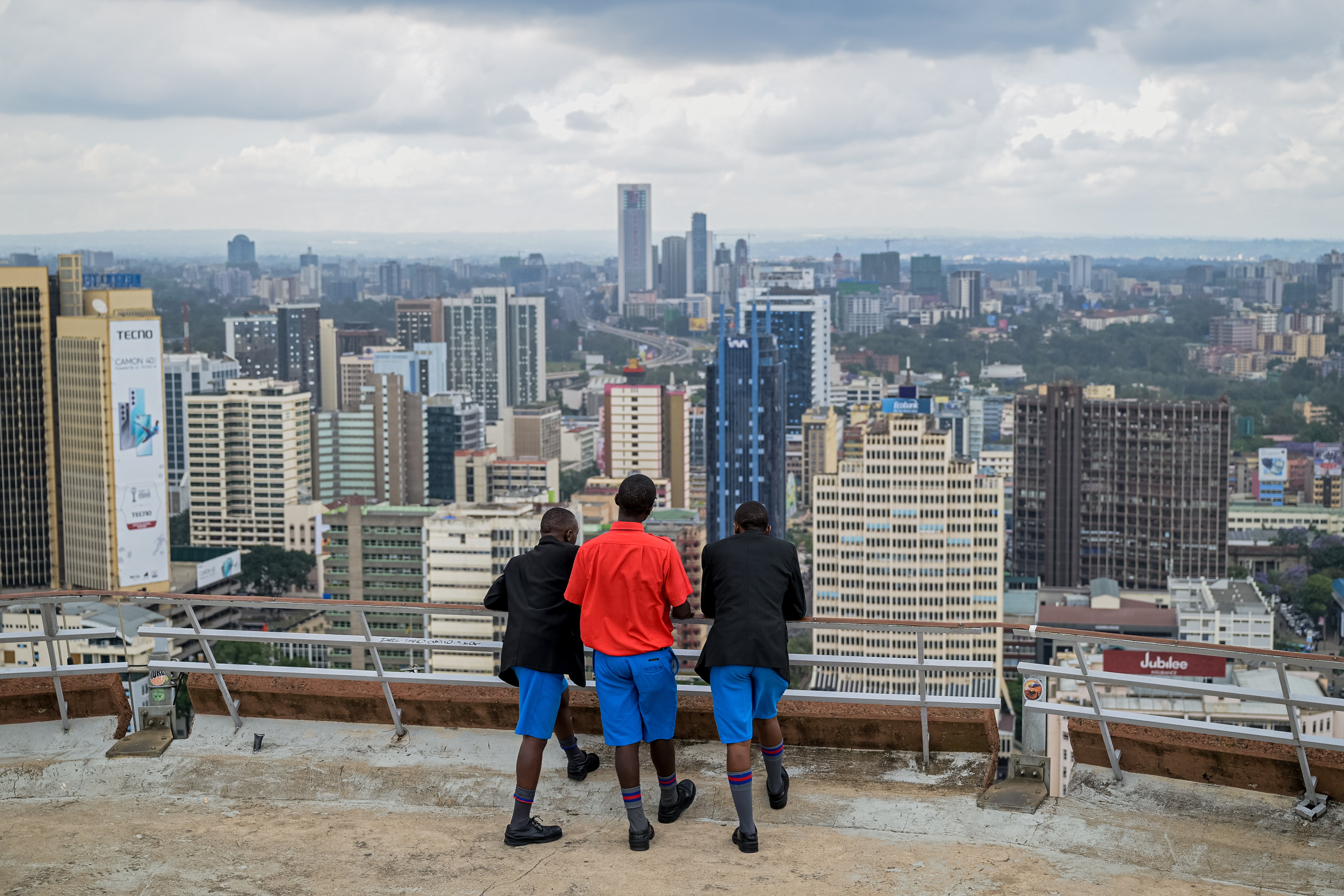 Tre unge gutter i skoleuniform står på en takterrasse og ser utover en storby med mange høye bygninger.
