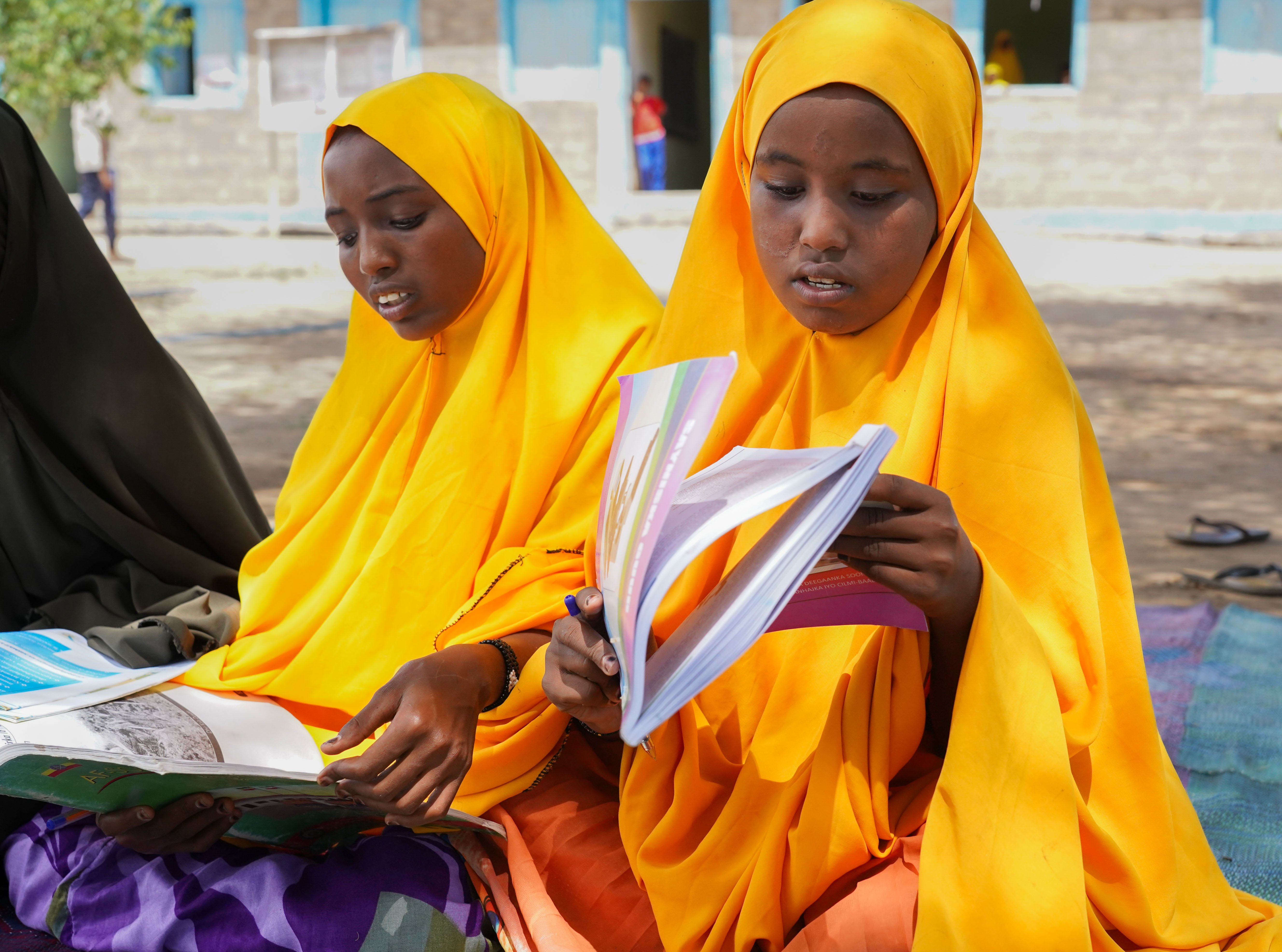 Two girls in yellow hijabs sit outdoors reading schoolbooks.