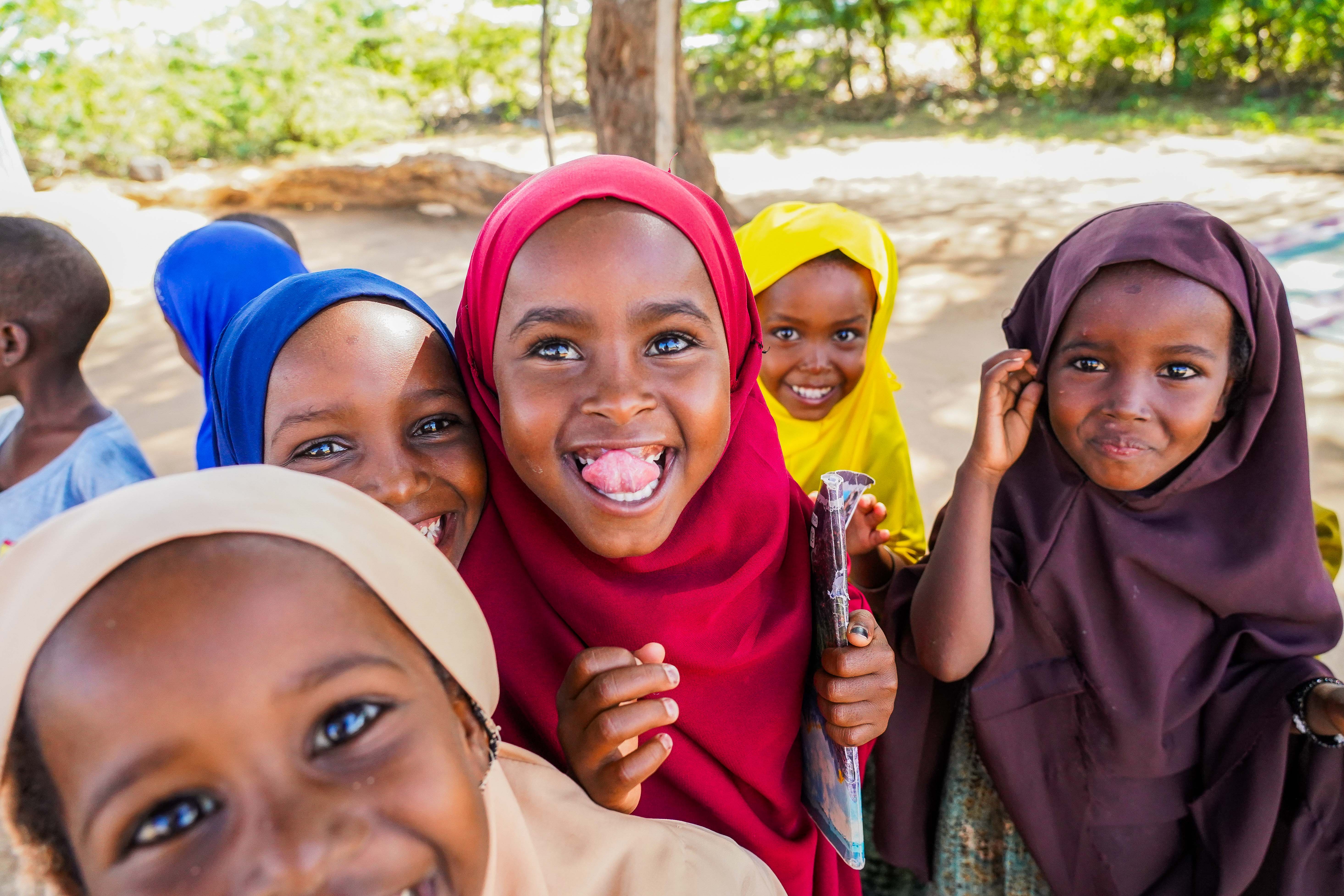 A group of young girls are standing together outdoors, wearing colourful headscarves and brightly coloured clothing.
